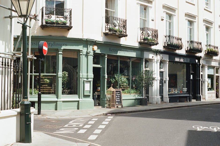 A storefront of a florist shop with a pale green facade and large glass windows displaying floral arrangements inside. The shop is situated on a corner of a quiet street with a pedestrian crossing nea