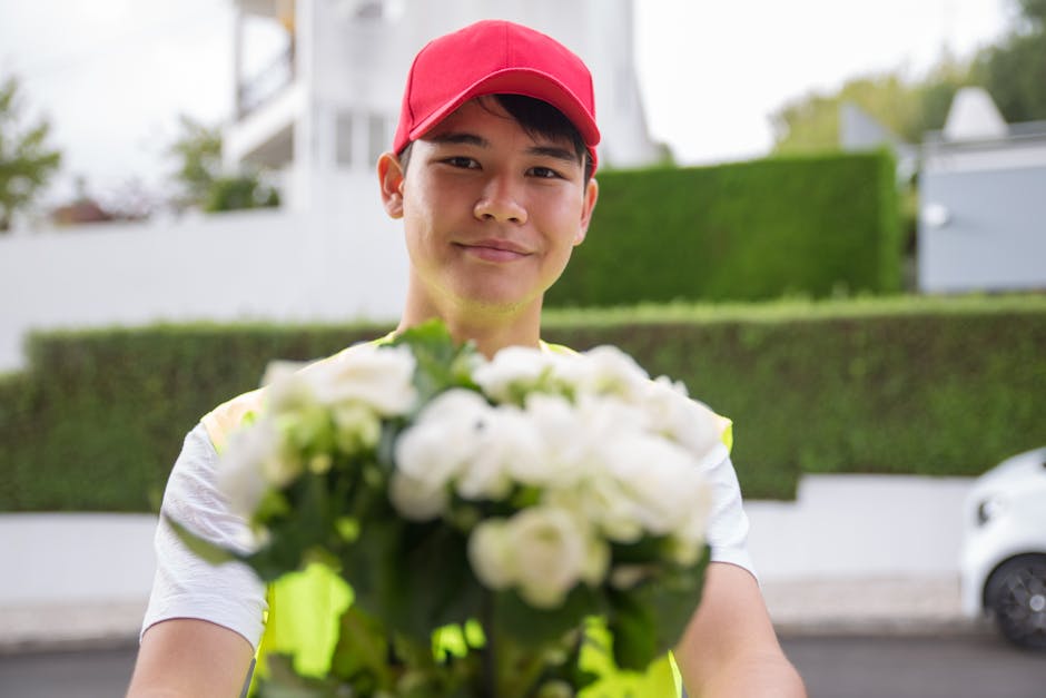 A delivery person wearing a red cap and a light-colored shirt is holding a bouquet of fresh white and green flowers, wrapped in clear floral wrapping with a green ribbon, in an outdoor suburban settin