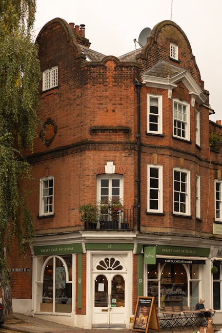 A charming red brick building with white-framed sash windows and decorative architectural details, housing a flower shop on the ground floor, illuminated by natural daylight. In front, a hand is holdi