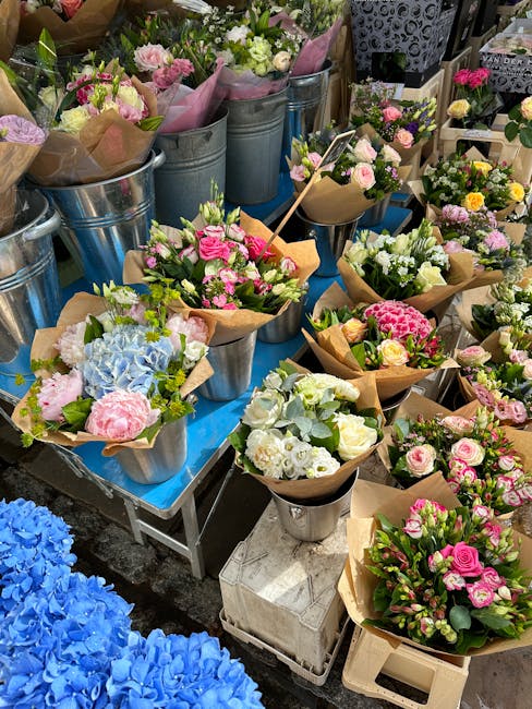A diverse selection of fresh floral arrangements displayed on a table, featuring various types of flowers including pink roses, white and pink lisianthus, blue hydrangeas, and assorted greenery. The b