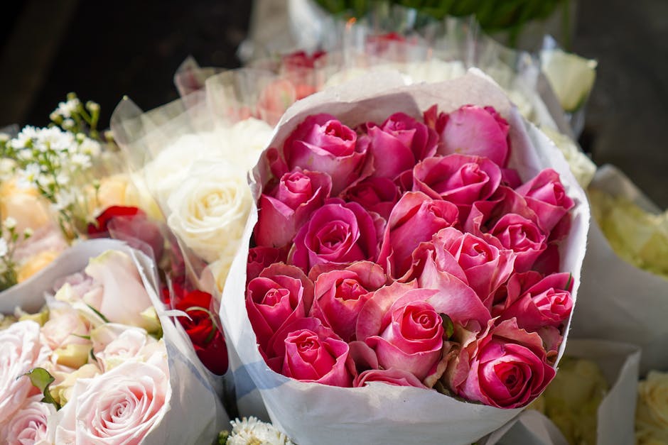 A bouquet of fresh pink roses with tightly curled petals and subtle white streaks, arranged in a lush, dense cluster. The roses are wrapped in white paper to enhance their vibrant colour and are posit