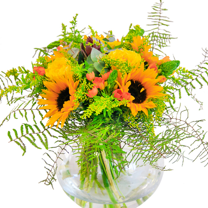 This exuberant floral composition, inspired by Dartmouth Park's serene atmosphere, bursts with colour and vitality. Large, glowing sunflowers and sumptuous yellow roses stand side by side, their petals layered in exuberant waves, spilling golden warmth across the bouquet. Deep green fern fronds tumble gracefully beyond the blooms, mingling with delicate sprays of solidago, while sprigs of slender green spikes add height and dimension. Glossy red hypericum berries, expertly scattered throughout, contrast against the matte petals-tiny jewels nestled in a sea of verdant foliage. Touches of bluish eucalyptus round out the palette, introducing a refreshing, subtle coolness that softens the whole. Meticulously arranged in a clear, globe-like vase, the bouquet's stems are revealed in shimmering water, reflecting the clean lines and bright interiors found in homes overlooking Hampstead Heath. Up close, one can sense the intricate texture: the plush sunflower faces, tightly furled rose blooms, the crisp texture of new greenery. This bouquet radiates celebration, perfect as a gift for milestone birthdays, heartfelt congratulations, or simply infusing a home in Dartmouth Park with seasonal joy and a flourish of craftsmanship.